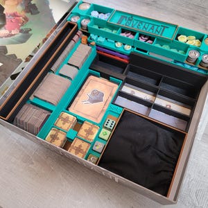 Puede incluir: Un organizador de juegos de mesa con compartimentos turquesa y negros. El organizador contiene cartas, fichas, dados y otros componentes del juego. La caja del juego está abierta, revelando el interior organizado. La palabra "COVENANT" es visible.