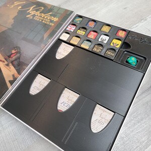 Puede incluir: Una caja de juego negra con una etiqueta blanca que dice "A Napoleon Designed by Ted Raicer". La caja está abierta y revela una bandeja negra con compartimentos para las piezas del juego y un dado verde.