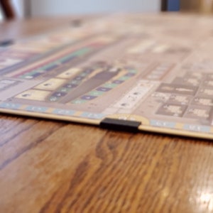 May include: A beige board game with a brown ship and blue and green squares. The board game is on a wooden table.