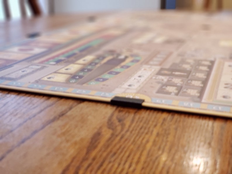 May include: A beige board game with a brown ship and blue and green squares. The board game is on a wooden table.