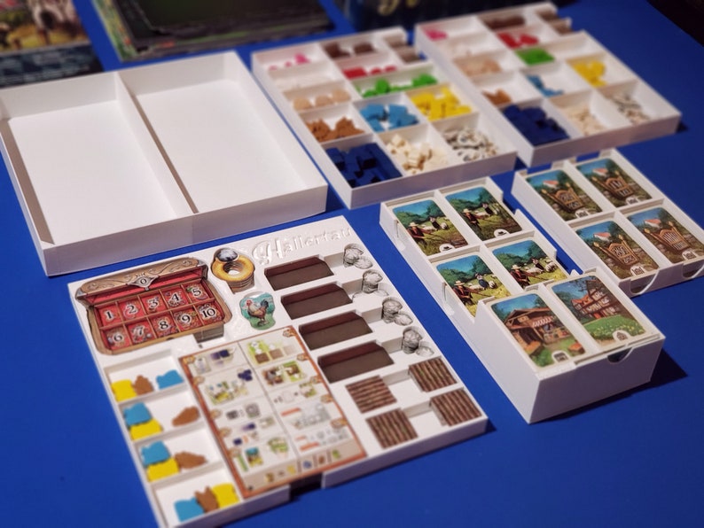 May include: A board game organizer with white trays and compartments filled with game pieces. The organizer includes card holders and spaces for tokens and other game components. The game board features the text "HallerTav".
