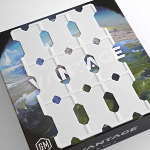 May include: A white plastic game board overlay with a grid pattern, placed over a game box. The box features a landscape image with the word "VANTAGE" in large white letters. The game box is black.