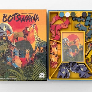 Puede incluir: Juego de mesa "Botswana" con una caja colorida que presenta ilustraciones de fauna africana. La caja abierta revela piezas de juego en una bandeja amarilla, incluyendo figuras de animales y fichas de madera. El tema del juego es un safari africano.