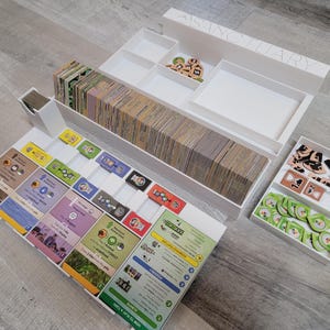 May include: White game organizer with multiple compartments and trays. Contains stacks of cards, wooden tokens, and game components. The word "SANCTUARY" is visible on the organizer.