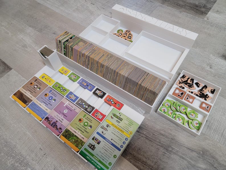 May include: White game organizer with multiple compartments and trays. Contains stacks of cards, wooden tokens, and game components. The word "SANCTUARY" is visible on the organizer.