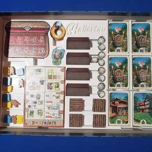 May include: A board game set with various components, including cards, wooden pieces, and tokens. The game box contains a chest with numbered slots, house-shaped pieces, and cards depicting gates and buildings. The game is titled "Hallertau".