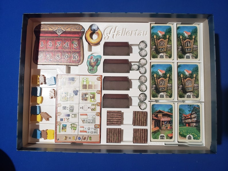 May include: A board game set with various components, including cards, wooden pieces, and tokens. The game box contains a chest with numbered slots, house-shaped pieces, and cards depicting gates and buildings. The game is titled "Hallertau".