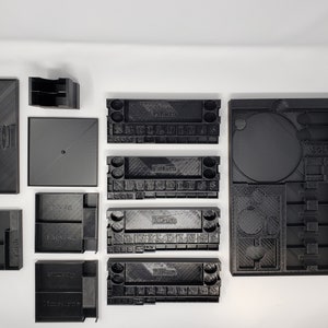 May include: A set of 10 black plastic storage containers with various compartments and labels. The containers are designed to organize and store small items, such as dice, tokens, and cards.