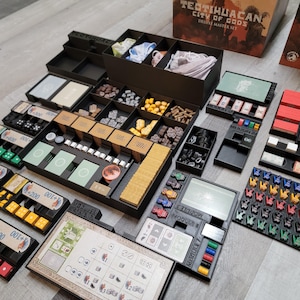 Può includere: Un organizzatore di gioco Teotihuacan: City of Gods Deluxe Master Set con più scomparti per riporre i pezzi del gioco. L'organizzatore è in plastica nera e ha una varietà di scomparti per riporre diversi tipi di pezzi del gioco, tra cui dadi, carte e gettoni. L'organizzatore include anche una serie di etichette che possono essere utilizzate per identificare il contenuto di ogni scomparto.