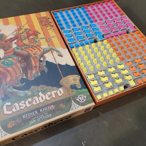 May include: A board game box with a colorful illustration of a knight riding a horse. The box is titled "Cascader" and features the name "Reiner Knizia" and "Artwork by Ian O'Toole". The game board is visible in the background, with colorful tiles arranged in a grid pattern.
