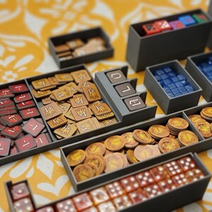 May include: A collection of board game components organized in gray trays. The trays hold red and tan tokens, blue and clear dice, and gold coins. The game pieces are arranged on a yellow patterned surface.