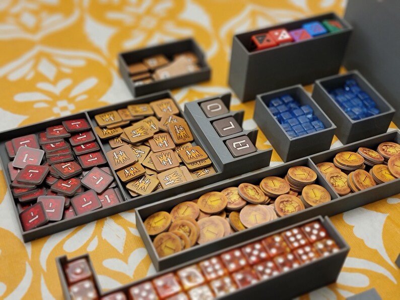 May include: A collection of board game components organized in gray trays. The trays hold red and tan tokens, blue and clear dice, and gold coins. The game pieces are arranged on a yellow patterned surface.