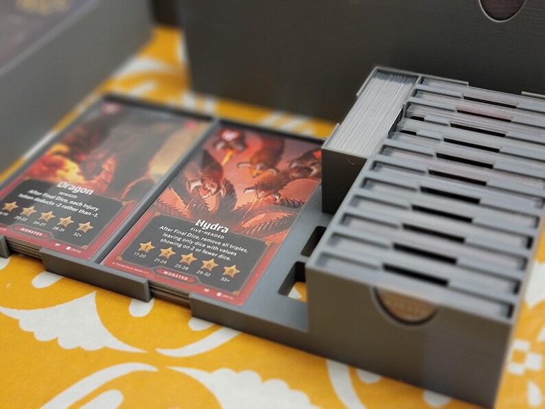 May include: A gray card game organizer with multiple compartments, holding stacks of cards. The cards feature illustrations of fantasy creatures, including a dragon and a hydra, with text describing game mechanics. The organizer is on a yellow patterned surface.