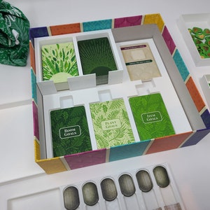 Puede incluir: Una caja de juego abierta con un inserto de espuma blanca. La caja tiene un dise&ntilde;o a rayas de colores con rayas verdes, amarillas, naranjas, rosas, azules y moradas. El inserto de espuma tiene compartimentos para cartas y fichas. Las cartas son verdes con ilustraciones de plantas y el texto "Room Goals", "Plant Goals" e "Item Goals". Las fichas son verdes y tienen forma de hojas.