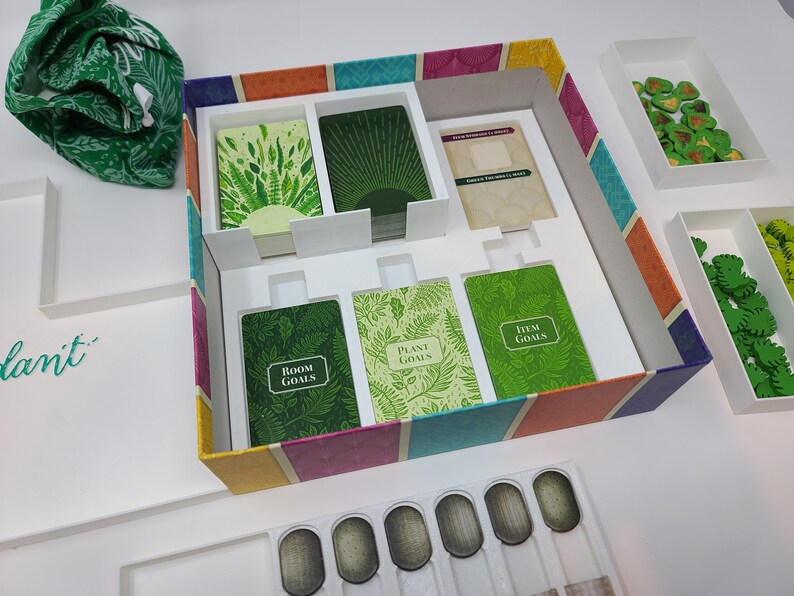 Puede incluir: Una caja de juego abierta con un inserto de espuma blanca. La caja tiene un dise&ntilde;o a rayas de colores con rayas verdes, amarillas, naranjas, rosas, azules y moradas. El inserto de espuma tiene compartimentos para cartas y fichas. Las cartas son verdes con ilustraciones de plantas y el texto "Room Goals", "Plant Goals" e "Item Goals". Las fichas son verdes y tienen forma de hojas.