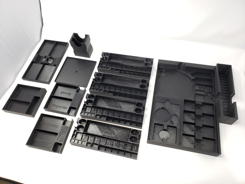 May include: A set of black plastic organizers for board games. The organizers are labeled with the names "Pillars", "Papyrus", "Bread", "Egusito", "Ankh", and "Solo".