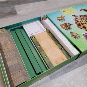 May include: Open board game box with the title "Sanctuary" visible on the box. The box is green and teal with images of animals. Inside the box are game pieces, including cards, green dividers, and wooden game boards.