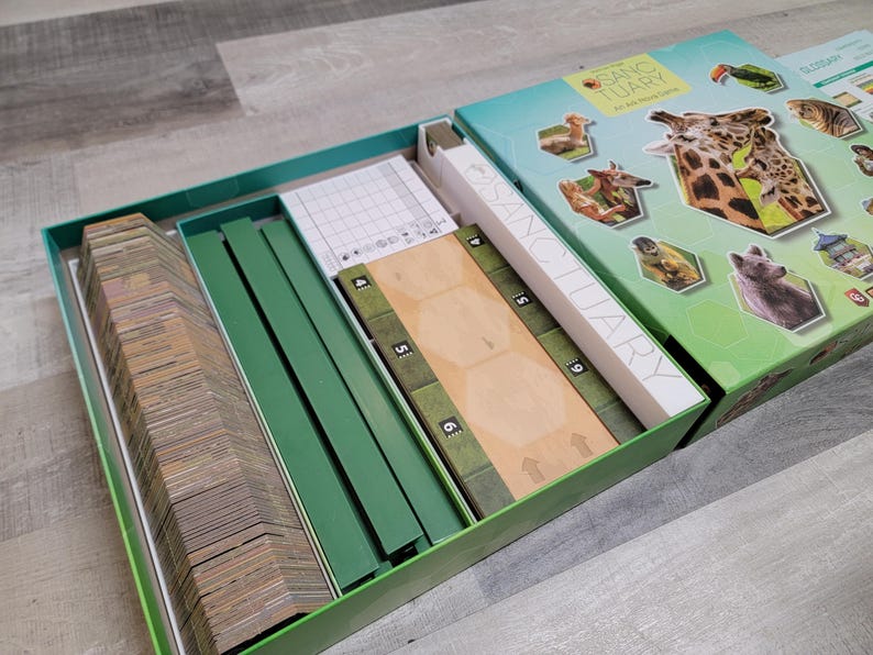 May include: Open board game box with the title "Sanctuary" visible on the box. The box is green and teal with images of animals. Inside the box are game pieces, including cards, green dividers, and wooden game boards.