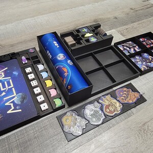 Peut inclure: Un jeu de soci&eacute;t&eacute; noir et bleu sur le th&egrave;me de l'espace. Le plateau de jeu pr&eacute;sente le mot "Whelm" en lettres dor&eacute;es. Le jeu comprend des d&eacute;s, des jetons et des cartes.