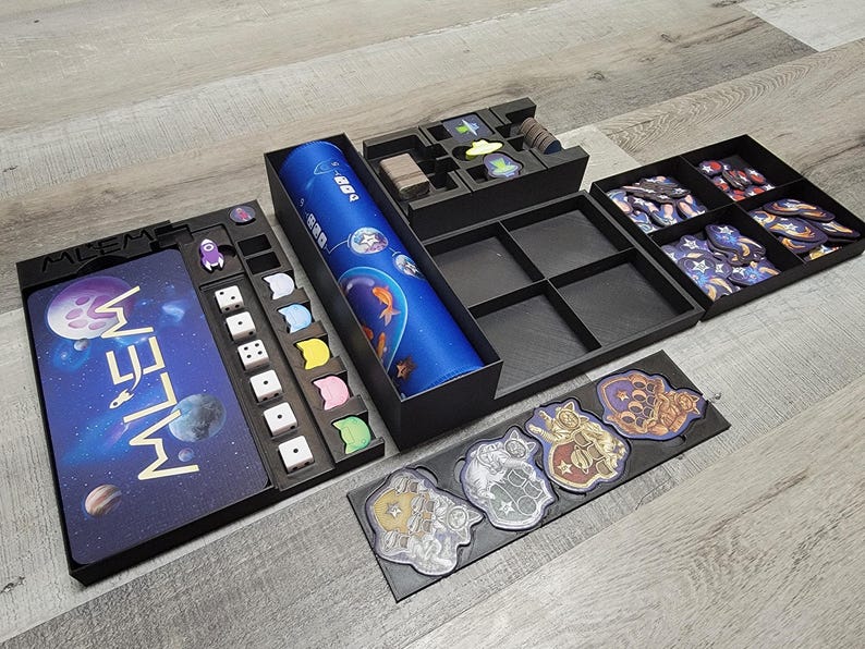 Peut inclure: Un jeu de soci&eacute;t&eacute; noir et bleu sur le th&egrave;me de l'espace. Le plateau de jeu pr&eacute;sente le mot "Whelm" en lettres dor&eacute;es. Le jeu comprend des d&eacute;s, des jetons et des cartes.