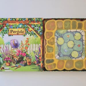 May include: A board game box and game pieces. The box features a garden scene with the word "Pergola". The game pieces show a pond with lily pads and a ladybug. The game is made by Rebel.