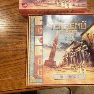 May include: A red and gold box with the title "Tekhenu: Obelisk of the Sun" and a picture of an ancient Egyptian scene. The box includes a rulebook and a game board with numbers 50-59.