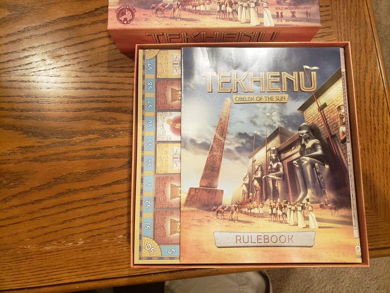 May include: A red and gold box with the title "Tekhenu: Obelisk of the Sun" and a picture of an ancient Egyptian scene. The box includes a rulebook and a game board with numbers 50-59.