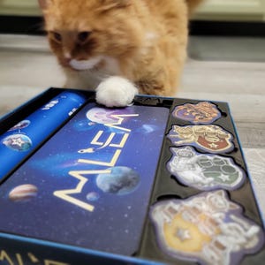 Peut inclure: Une bo&icirc;te de jeu de soci&eacute;t&eacute; bleue et dor&eacute;e avec le mot "MILEA" imprim&eacute; sur le devant. La bo&icirc;te est ouverte et contient plusieurs pi&egrave;ces de jeu, dont un plateau de jeu bleu et blanc, un sac en tissu bleu et plusieurs petits jetons color&eacute;s.