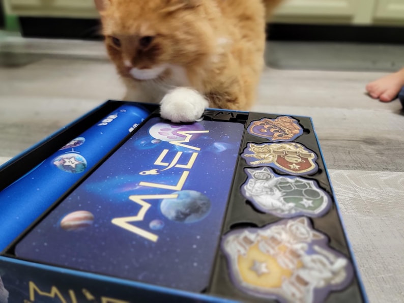 Peut inclure: Une bo&icirc;te de jeu de soci&eacute;t&eacute; bleue et dor&eacute;e avec le mot "MILEA" imprim&eacute; sur le devant. La bo&icirc;te est ouverte et contient plusieurs pi&egrave;ces de jeu, dont un plateau de jeu bleu et blanc, un sac en tissu bleu et plusieurs petits jetons color&eacute;s.