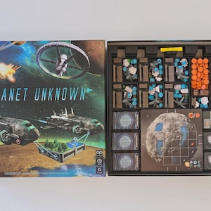 May include: Open board game box titled "Planet Unknown" with a space theme. The box art features spaceships, a space station, and a planet. Inside, the game components are organized in compartments, including game pieces and cards.
