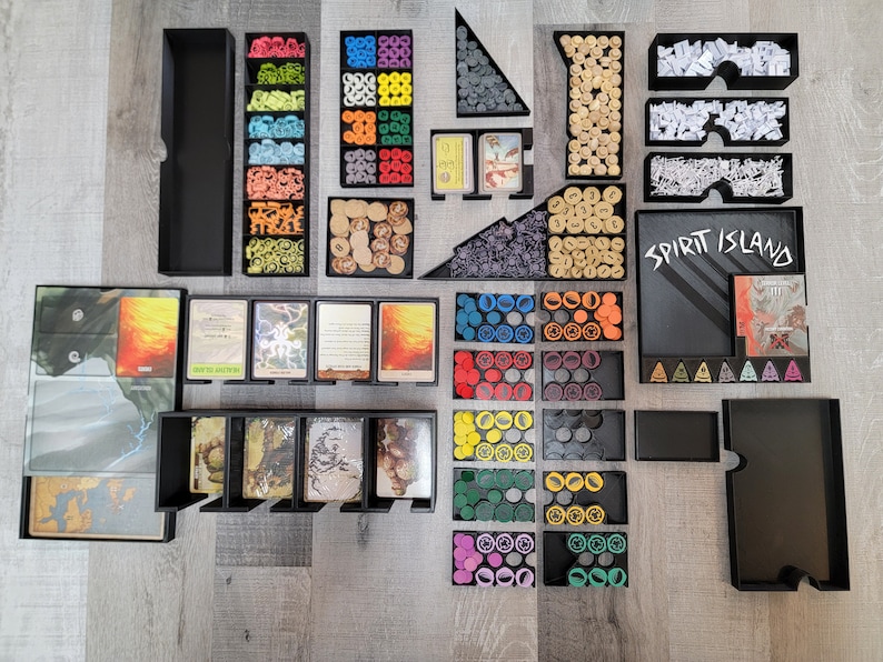 May include: A Spirit Island board game organiser with multiple compartments for storing game components. The organiser includes compartments for cards, tokens, and other game pieces. The organiser is made of black plastic and has a sleek, modern design.