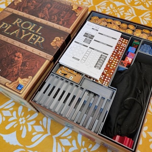 May include: Open board game box with the title "ROLL PLAYER" in gold lettering. The box contains game components like dice, cards, tokens, and a rule sheet. The box has a brown and gold design with fantasy artwork.