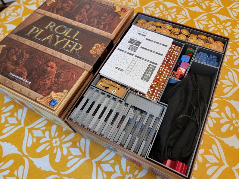 May include: Open board game box with the title "ROLL PLAYER" in gold lettering. The box contains game components like dice, cards, tokens, and a rule sheet. The box has a brown and gold design with fantasy artwork.
