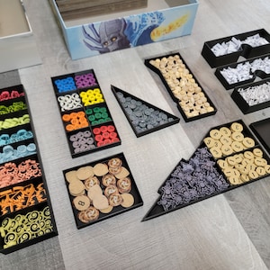 May include: A black organiser with multiple compartments filled with various coloured game pieces, including small round tokens, cubes, and stars. The organiser is designed to store and organise game components.