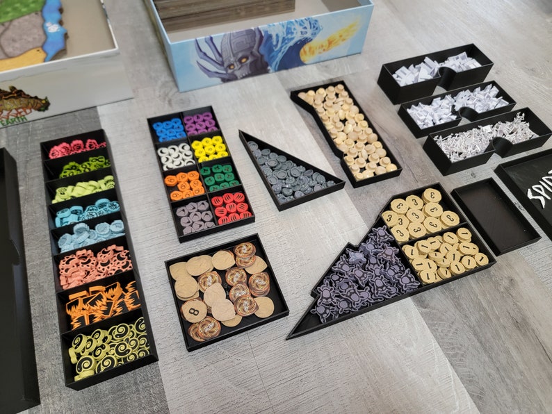 May include: A black organiser with multiple compartments filled with various coloured game pieces, including small round tokens, cubes, and stars. The organiser is designed to store and organise game components.