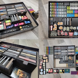 May include: A black and gray board game organizer with multiple compartments for storing game pieces, cards, and other components. The organizer is labeled "Hegemony".