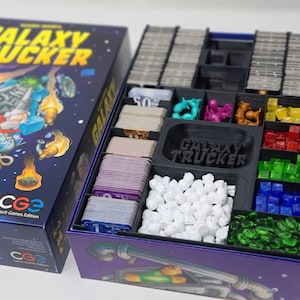 May include: A purple and black board game box with a galaxy trucker theme. The box is open and shows a black plastic insert with compartments for game pieces, cards, and dice. The insert has the words "Galaxy Trucker" printed on it.