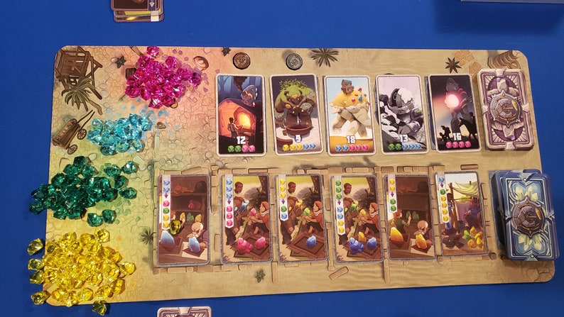 Peut inclure: Un plateau de jeu avec des gemmes color&eacute;es, des cartes et divers personnages. Le plateau est en bois et a une couleur brun clair. Les cartes sont de tailles et de formes diff&eacute;rentes, et elles ont des images diff&eacute;rentes. Les gemmes sont de couleurs diff&eacute;rentes et sont dispos&eacute;es en pile au centre du plateau.