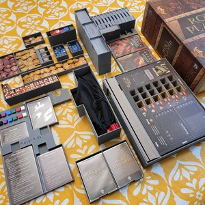 May include: An open board game box with various components, including dice, tokens, cards, and player aids. The box is brown with the words "ROLL PLAYER" on it. The game is set on a yellow patterned surface.