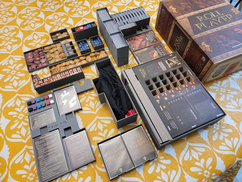 May include: An open board game box with various components, including dice, tokens, cards, and player aids. The box is brown with the words "ROLL PLAYER" on it. The game is set on a yellow patterned surface.