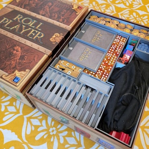 May include: Open board game box with the title "ROLL PLAYER" on the lid. The box is brown with gold accents and contains various game components, including cards, dice, and tokens. The game is set up in a yellow floral pattern.