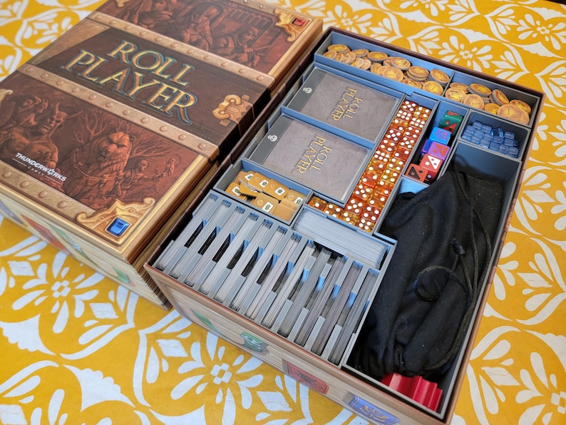 May include: Open board game box with the title "ROLL PLAYER" on the lid. The box is brown with gold accents and contains various game components, including cards, dice, and tokens. The game is set up in a yellow floral pattern.