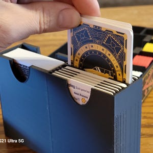May include: A black card game box with a white card being inserted. The card has a gold and black design with the text "Heist Process" and "Play Each game has..."