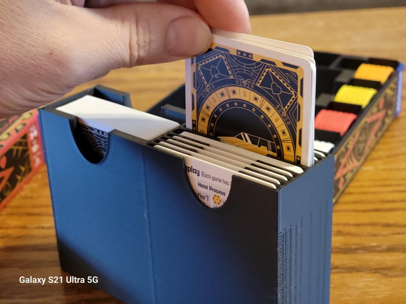 May include: A black card game box with a white card being inserted. The card has a gold and black design with the text "Heist Process" and "Play Each game has..."