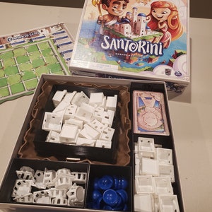 May include: A white cardboard box with a custom insert for organising the Santorini board game pieces. The insert has compartments for the game's white buildings, blue player pieces, and cards. The box lid features a colourful illustration of the game's characters and the game's name, Santorini.