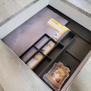 May include: A black, compartmentalized game box insert with a gray and white interior. The insert holds game cards and components, including cards labeled "ACTIONS" and a decorative game piece. The box is set on a light gray wooden surface.