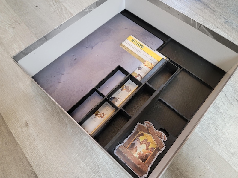 May include: A black, compartmentalized game box insert with a gray and white interior. The insert holds game cards and components, including cards labeled "ACTIONS" and a decorative game piece. The box is set on a light gray wooden surface.