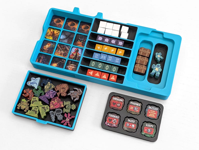 Może przedstawiać: Turkusowy organizer do gier planszowych z wieloma przegr&oacute;dkami na elementy gry. Organizer zawiera miejsca na karty, żetony i miniaturowe figurki. Dodatkowe tace zawierają elementy gry i żetony z efektami statusu, takimi jak "Enraging" i "Sleepy".