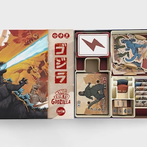 May include: A board game box set featuring Godzilla artwork on the lid with Japanese text and the title King of Tokyo Godzilla. The open box contains organized plastic trays holding card decks, cardboard character standees, and wooden dice.