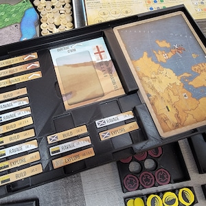 May include: A black board game tray with various coloured tokens and cards. The tray has slots for different game components, including a map, cards, and tokens. The tray is labelled with the words "Build", "Ravage", and "Explore".